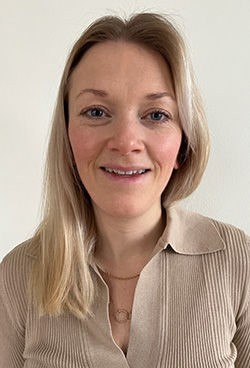 Kvinna med blont hår och beige tröja ler mot kameran framför ljus bakgrund.
