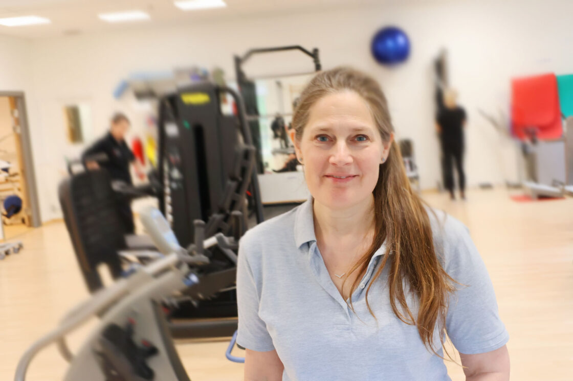 Kvinna med långt hår och ljusblå pikétröja i ett ljust gym med träningsmaskiner och suddiga personer i bakgrunden.