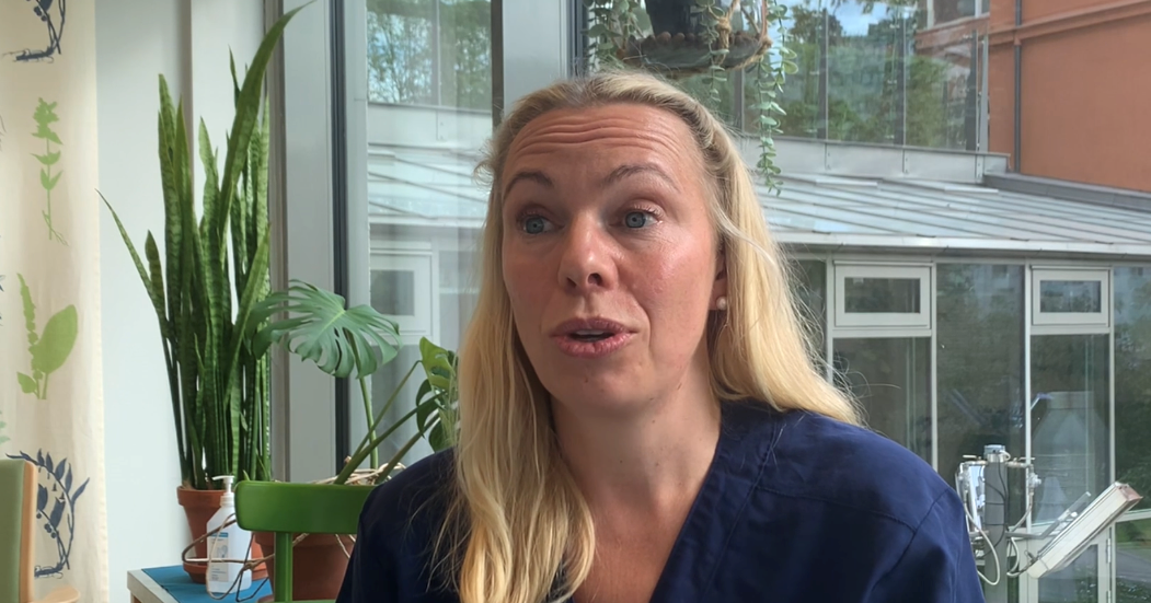 Kvinna med långt blont hår i blå tröja sitter inomhus med växter och stora fönster i bakgrunden.