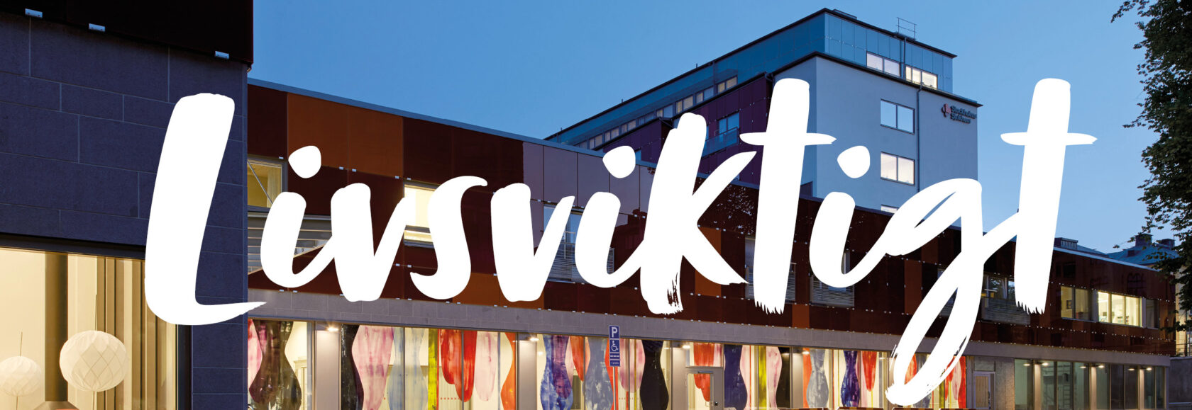 Texten "Livsviktigt" i vitt över en modern byggnad i kvällsljus.