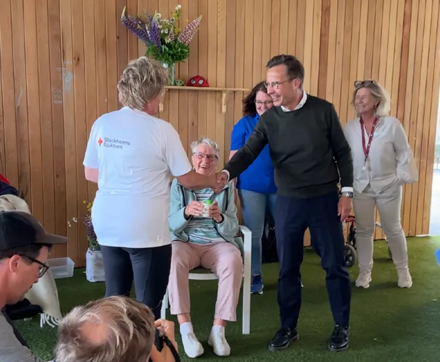En äldre kvinna sitter på en stol, omgiven av leende personer inomhus med träväggar och blommor i bakgrunden.