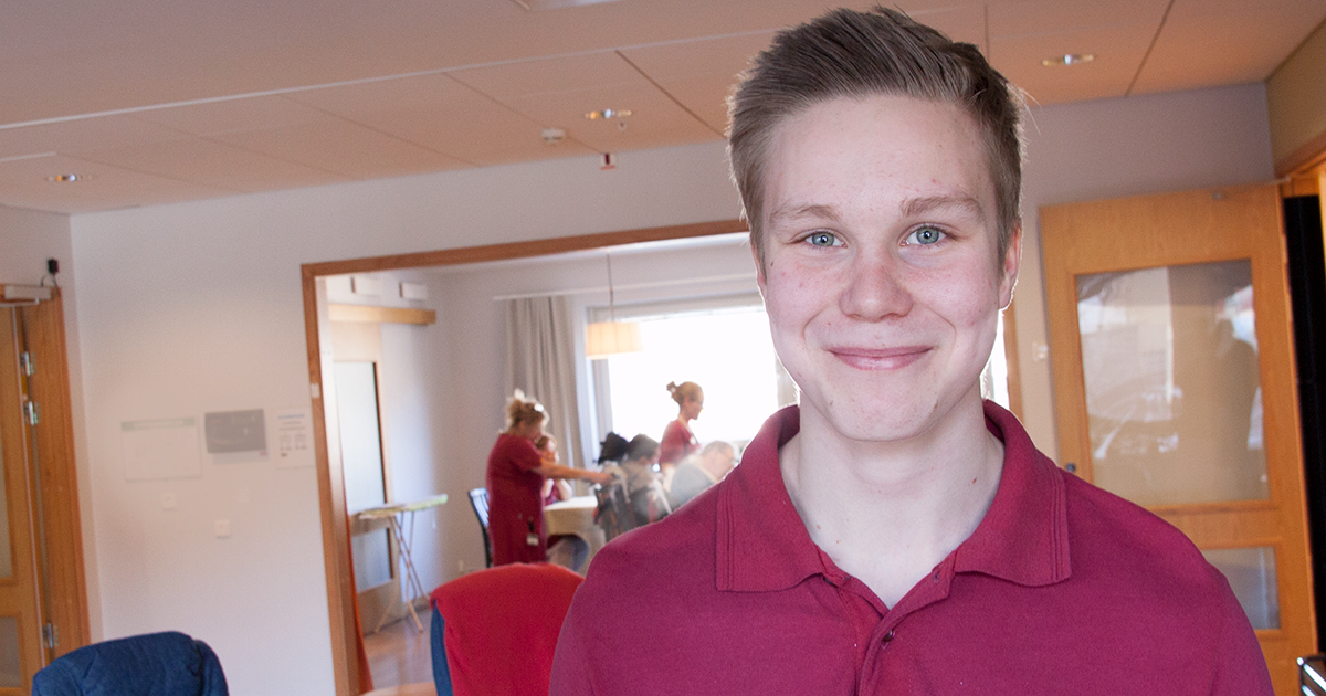 Ung man i röd pikétröja ler mot kameran, folk arbetar i bakgrunden i ett ljust rum.
