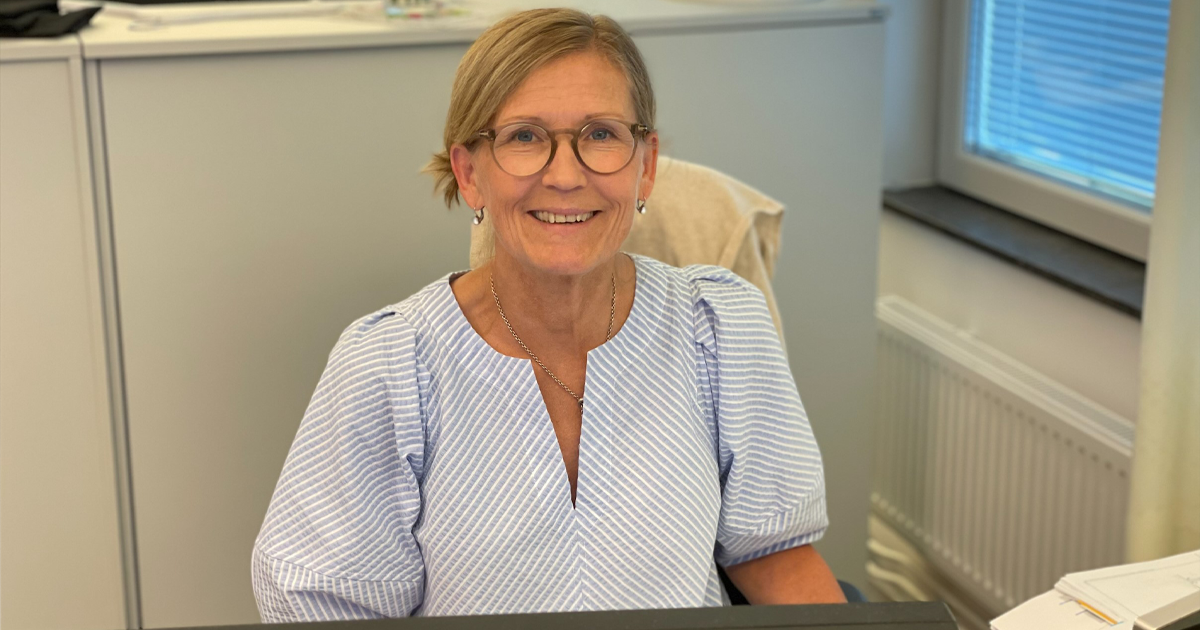 Leende kvinna med glasögon sitter vid skrivbord på kontor, iklädd ljusblå randig blus.