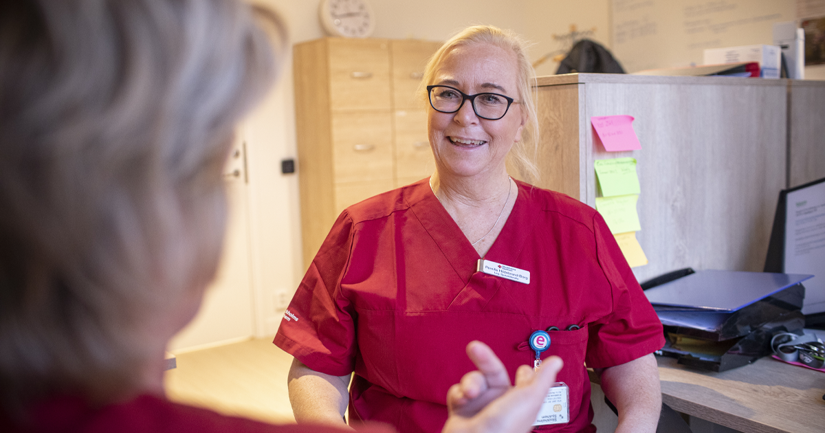 Sjuksköterska i röd uniform sitter och samtalar med en person på kontor med post-it-lappar på väggen.