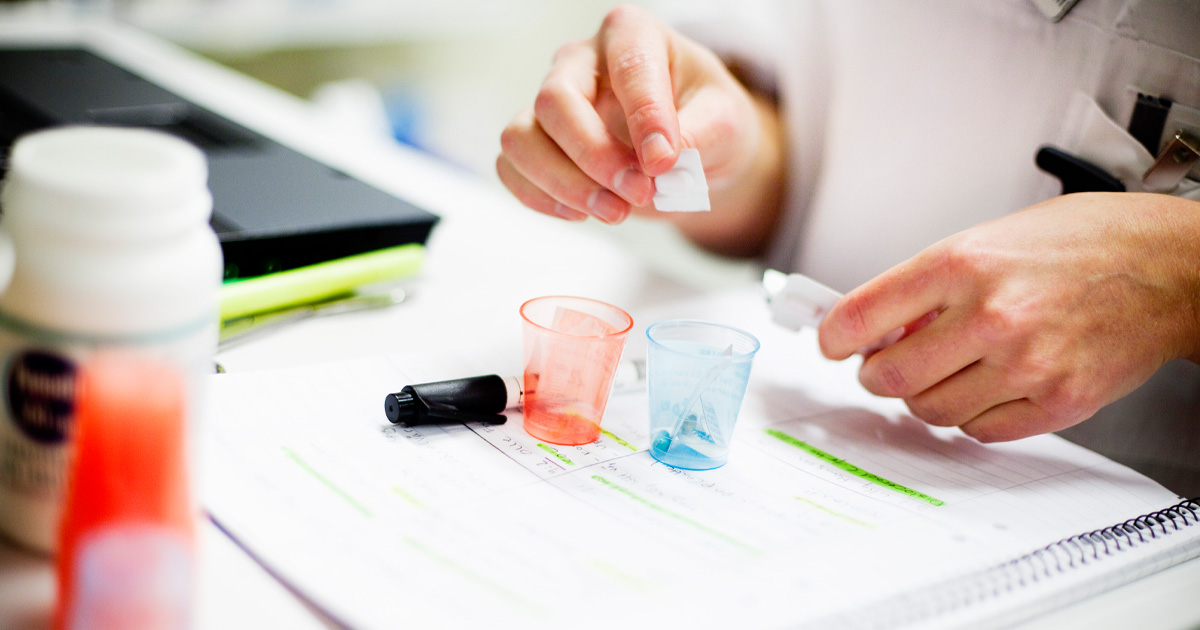 Person delar medicin i plastmuggar bredvid anteckningar och en flaska på ett skrivbord.
