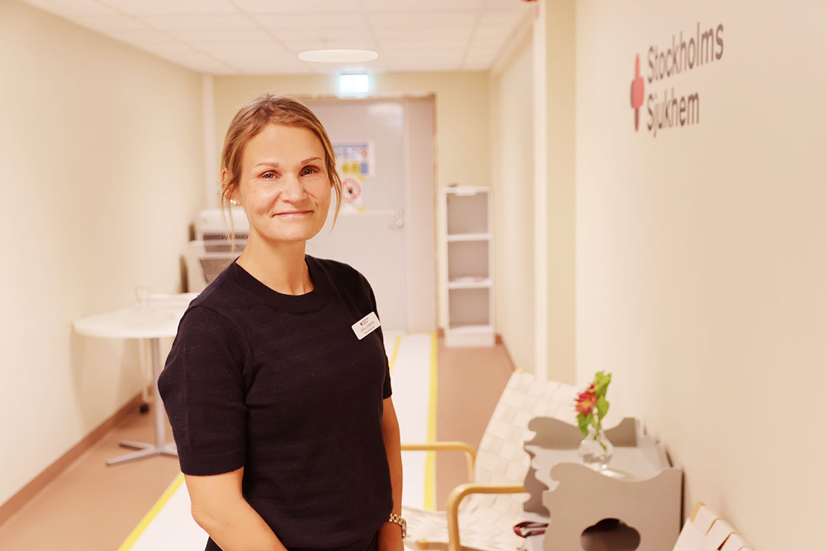 Kvinna i svart tröja står i en ljus korridor med texten "Stockholms Sjukhem" på väggen.