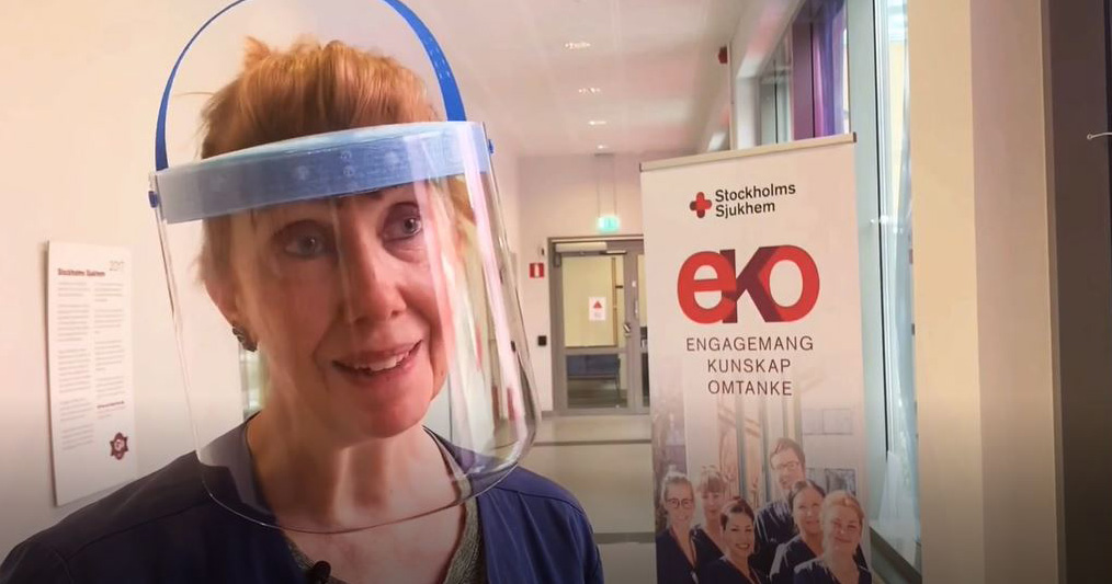 Kvinna med visir i sjukhuskorridor, skylt från Stockholms Sjukhem syns i bakgrunden.