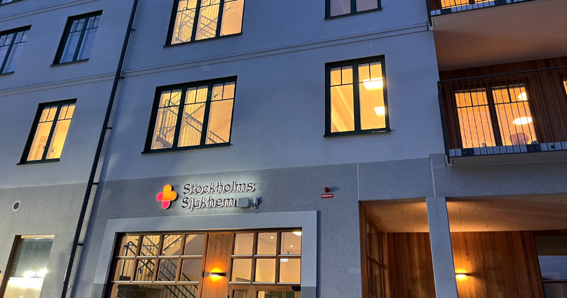 Fasad på Stockholms Sjukhems äldreboende Nyckelharpan i Upplands Väsby. Kvällsbild där huset har upplysta fönster och entré i kvällsljus.