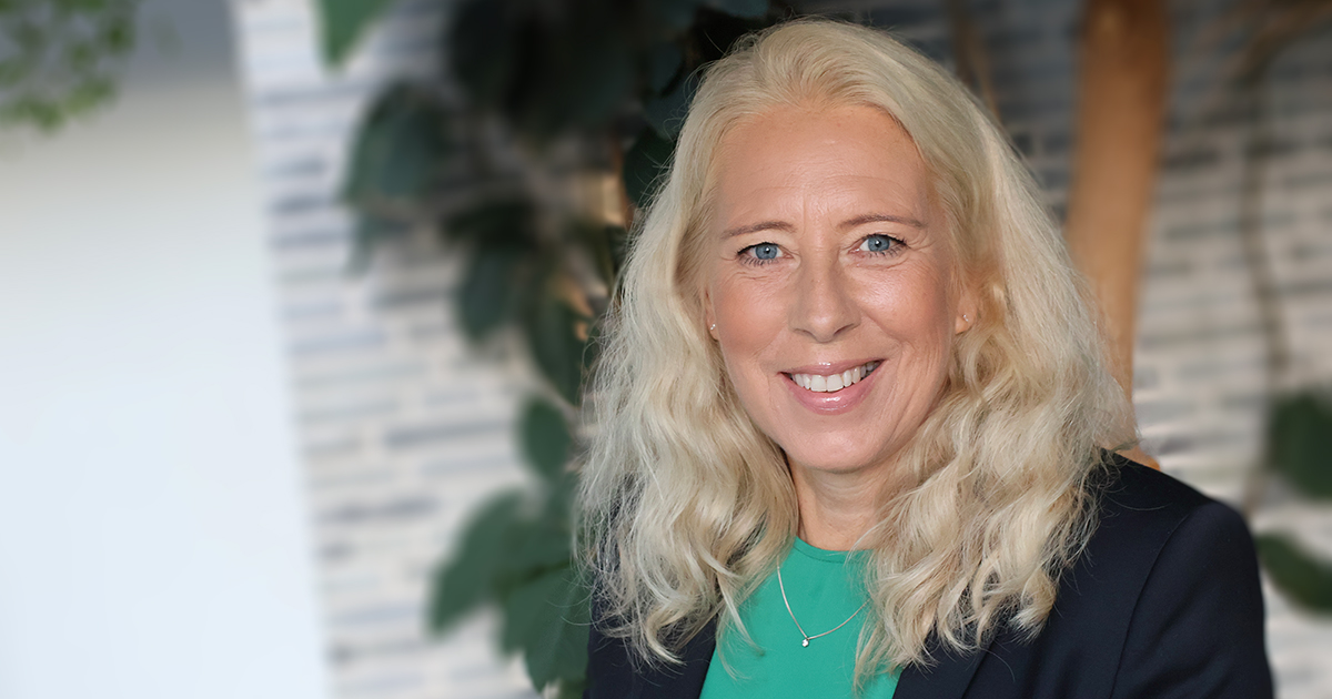 Kvinna med långt blont hår ler mot kameran, bär grön topp och svart kavaj, suddig bakgrund med växt.