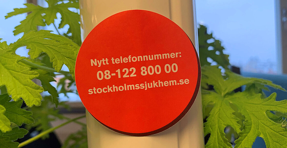 Röd rund lapp med text: Nytt telefonnummer 08-122 800 00 stockholmssjukhem.se, på vit yta med gröna blad.