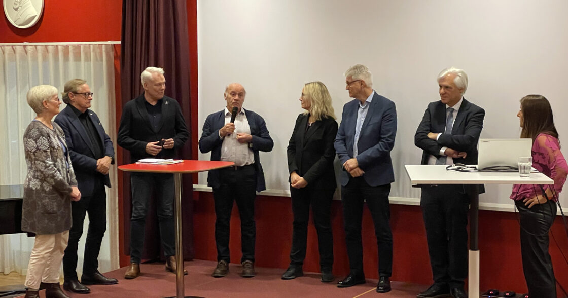 Sju personer står på en scen, en talar i mikrofon. Två bord och en laptop syns. Röd och vit bakgrund.