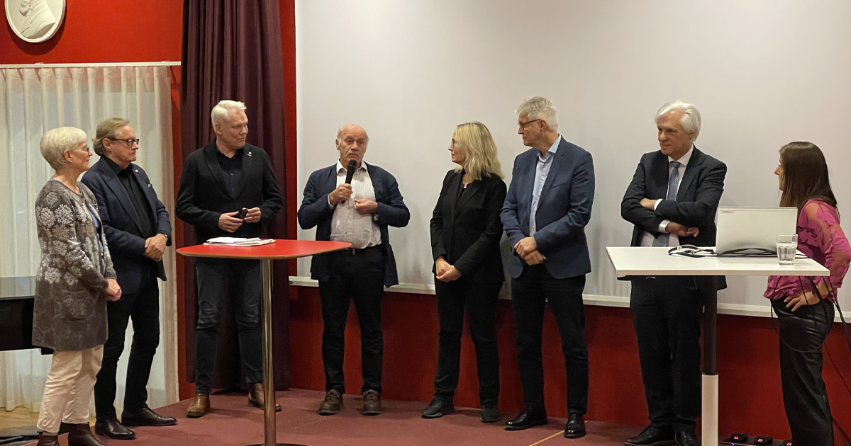 Sju personer står på en scen, en talar i mikrofon. Två bord och en laptop syns. Röd och vit bakgrund.