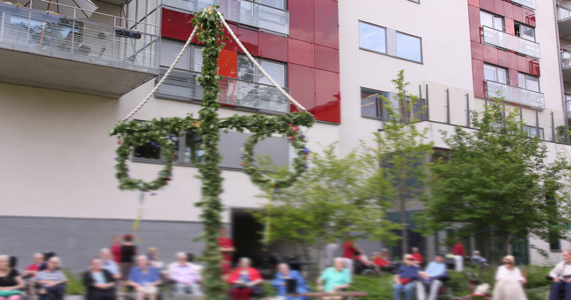 Midsommarstång framför ett modernt bostadshus med människor sittande i bakgrunden.