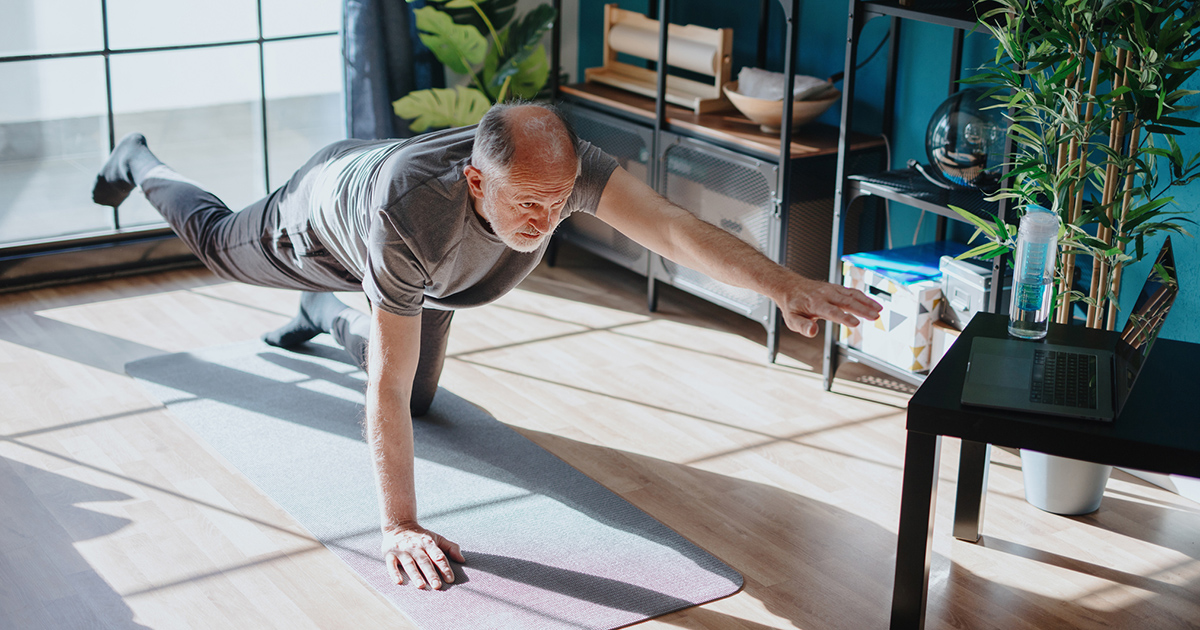 Äldre man tränar balansövning på yogamatta hemma, tittar på dator på bord, solljus genom fönster.