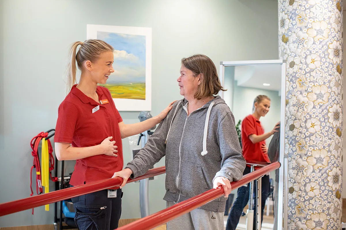 Medarbetare i arbetskläder ler och tittar på patient som gåtränar.