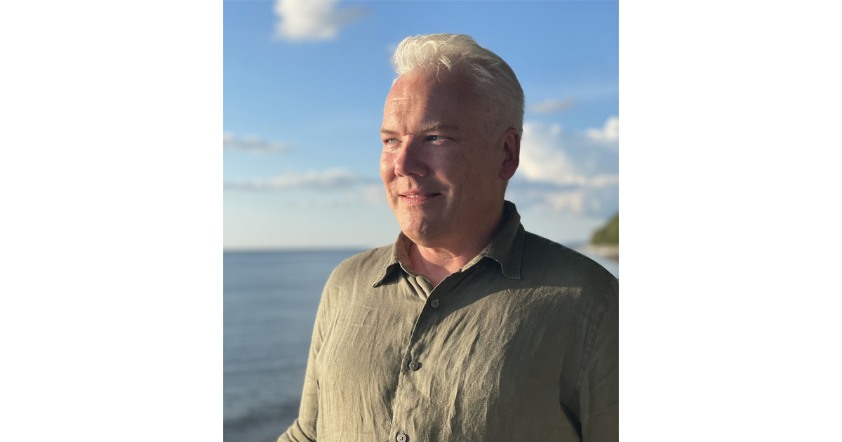 Man med ljust hår och skjorta står vid havet i solnedgång, moln och blå himmel i bakgrunden.