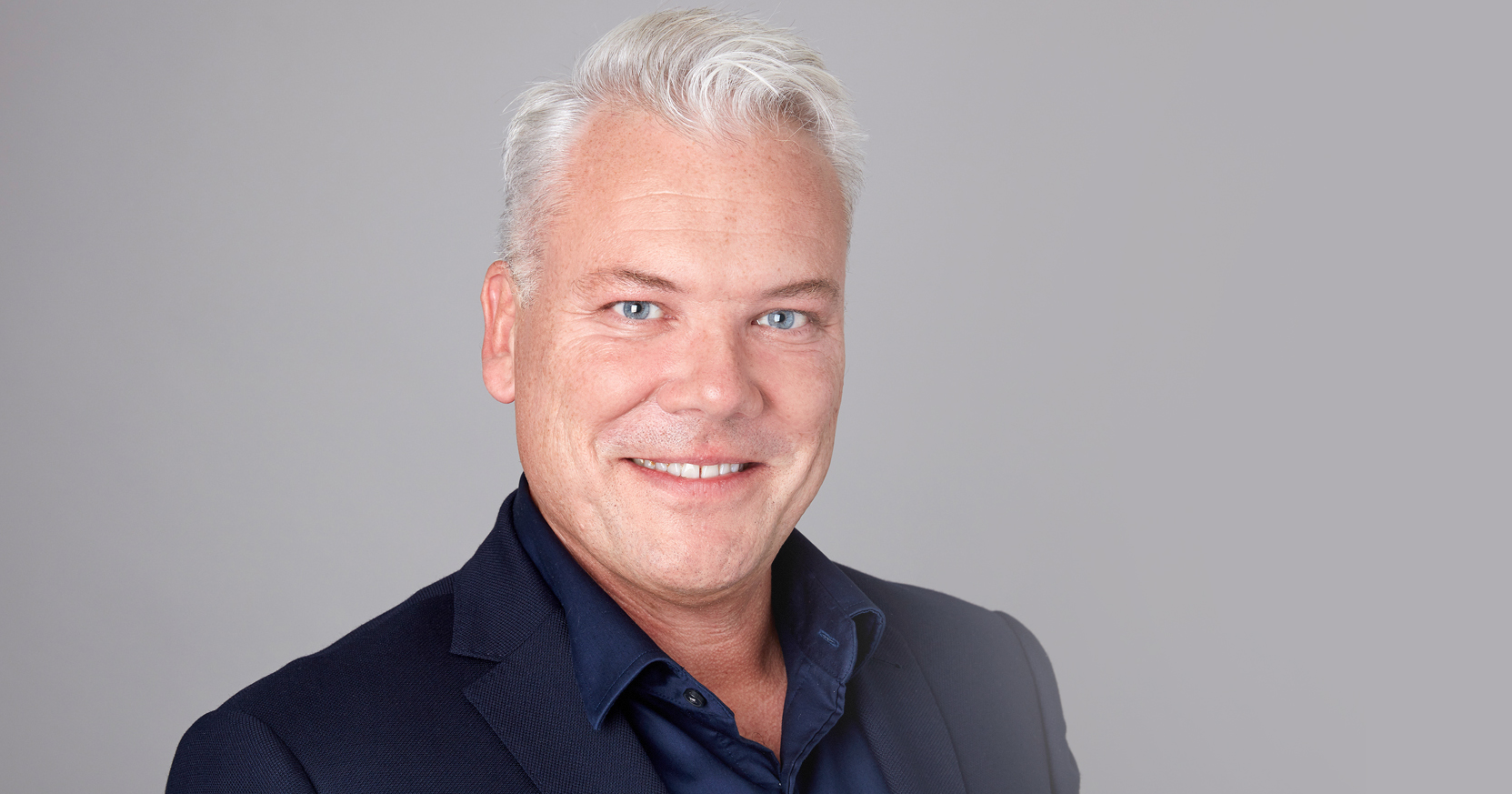 Leende man med grått hår i mörk kavaj mot ljus bakgrund.