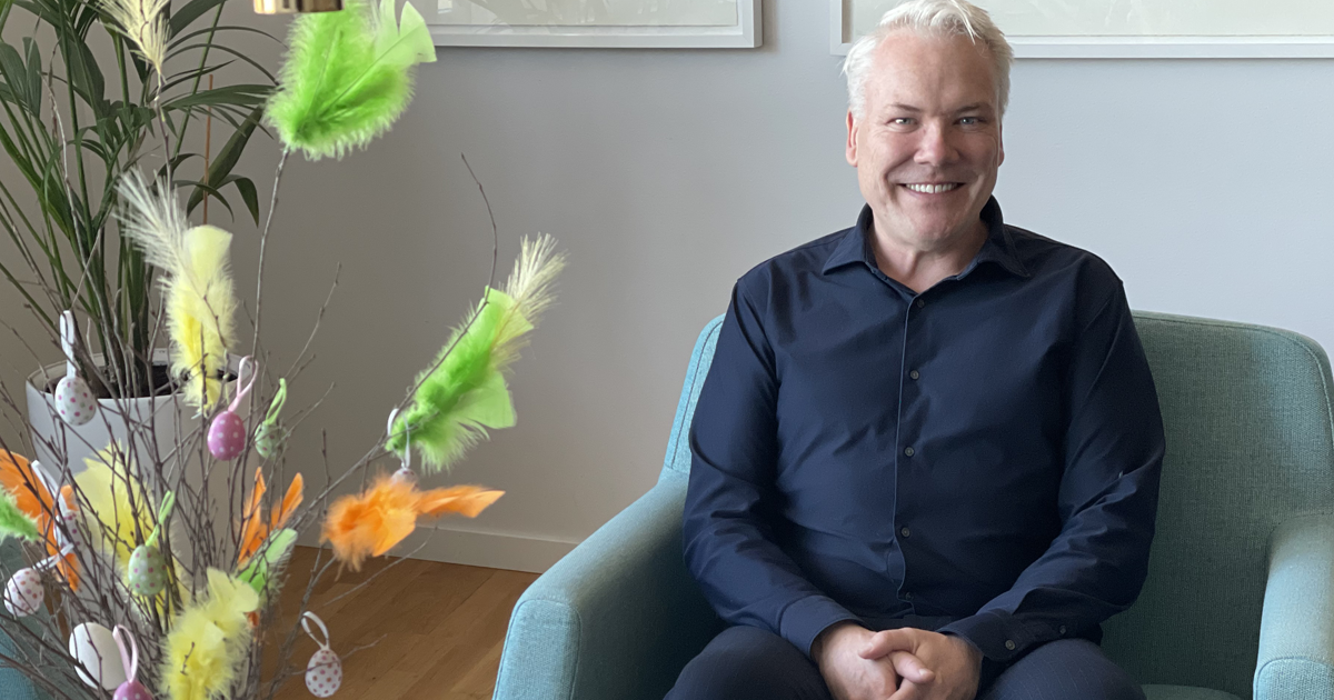 Man i blå skjorta sitter i fåtölj bredvid påskris med färgglada fjädrar och ägg, inomhusmiljö.