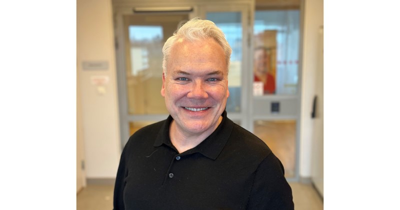 Leende man med grått hår och svart tröja står inomhus framför en glasdörr.