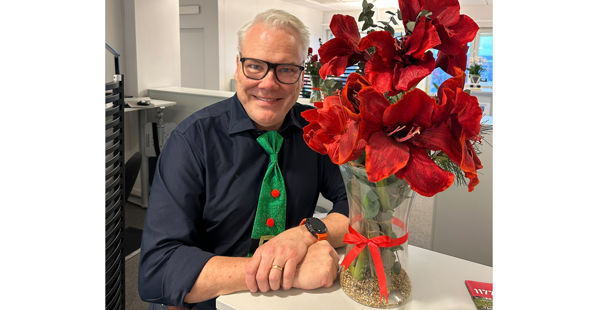 Peter Ceder, äldreomsorgschef, ler mot kameran. I klädd glasögon och grön slips. Bredvid en vas med stora röda blommor på ett kontor.