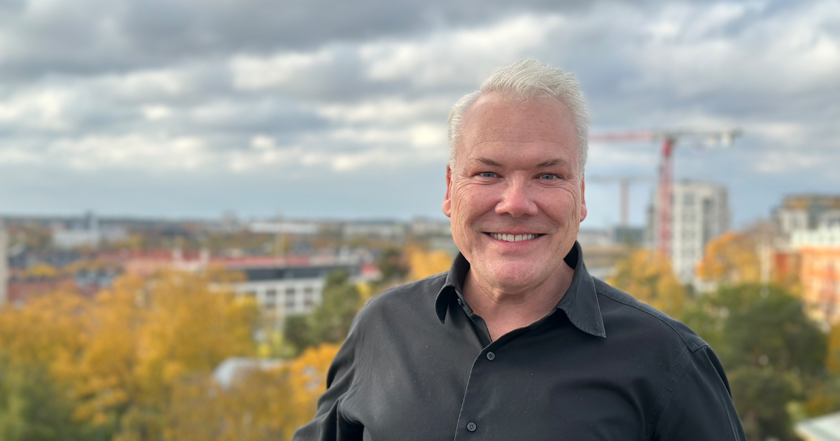 Leende man med vitblont hår i svart skjorta utomhus, suddig stadsmiljö och höstfärger i bakgrunden.