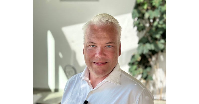 Man med ljust hår och vit skjorta står inomhus, suddig bakgrund med växter och ljusa skuggor.