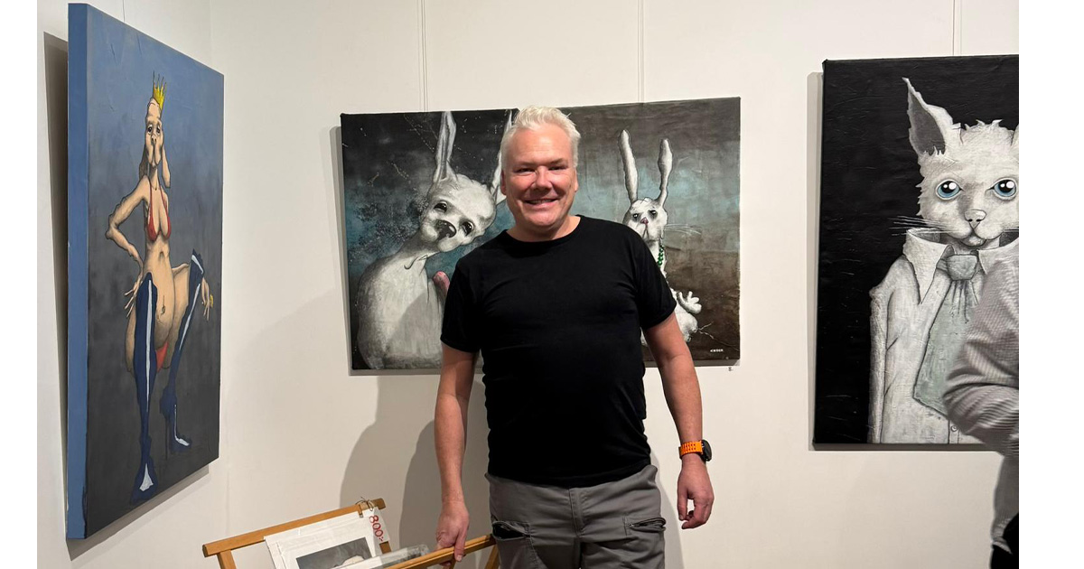 Leende man står framför surrealistiska målningar av djur och människor i ett konstgalleri.