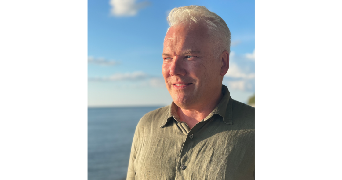 Man med grått hår och grön skjorta står utomhus nära vatten och ser leende mot solen.
