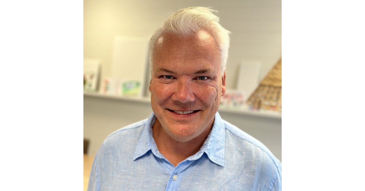 Leende man med vitt hår och blå skjorta står inomhus med suddig bakgrund.