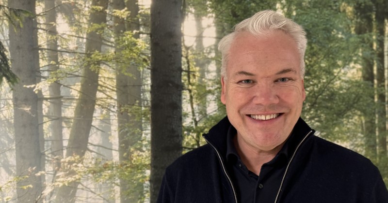 Leende man med ljust hår står framför en bakgrund av skog med solstrålar genom träden.