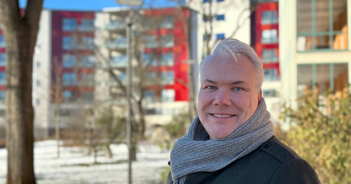 Leende man med grå scarf utomhus, bostadshus i bakgrunden, soligt vinterväder.