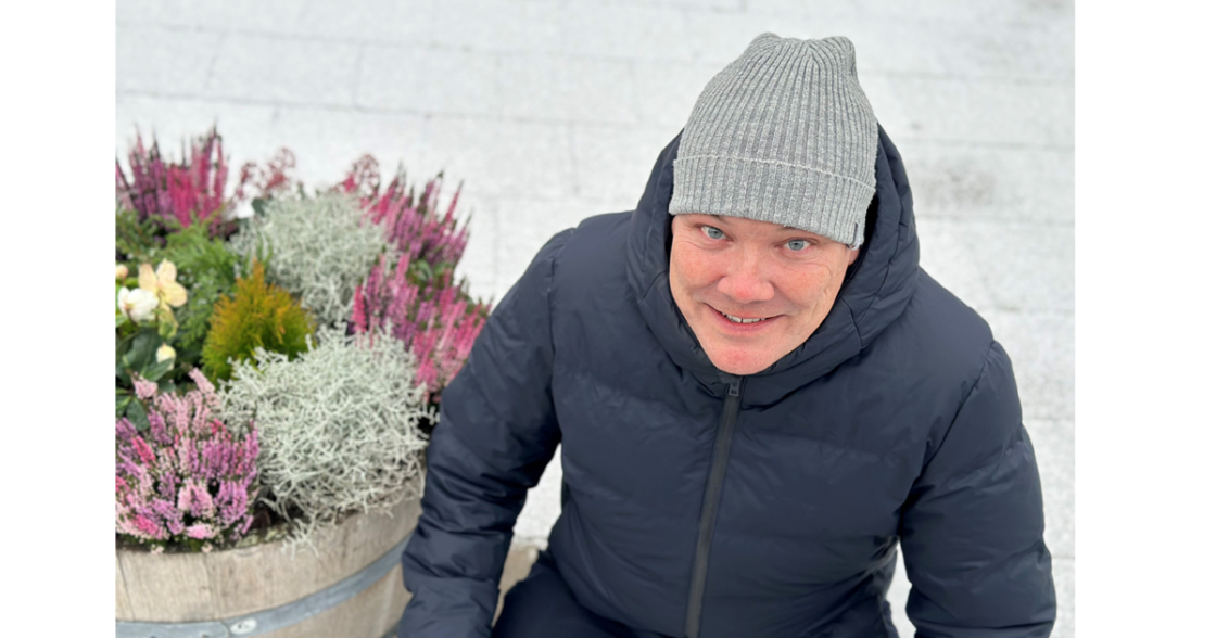 Man i grå mössa och mörk jacka sitter bredvid en blomkruka utomhus vintertid.