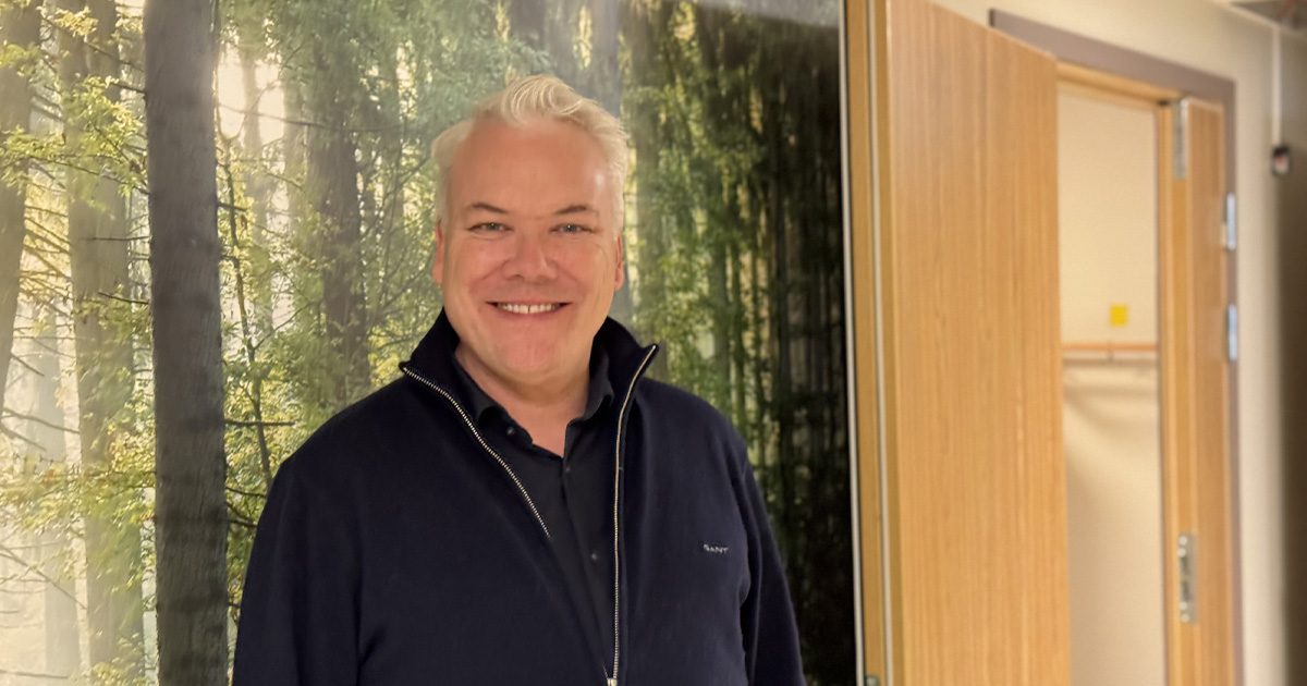 Leende man i mörk tröja står inomhus, med skogstapet och öppen dörr i bakgrunden.