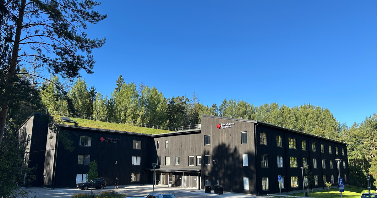 Äldreboendet Poesin i Nacka. Svart byggnad med grönt tak, Stockholms Sjukhem-logga, omgiven av skog under klarblå himmel.