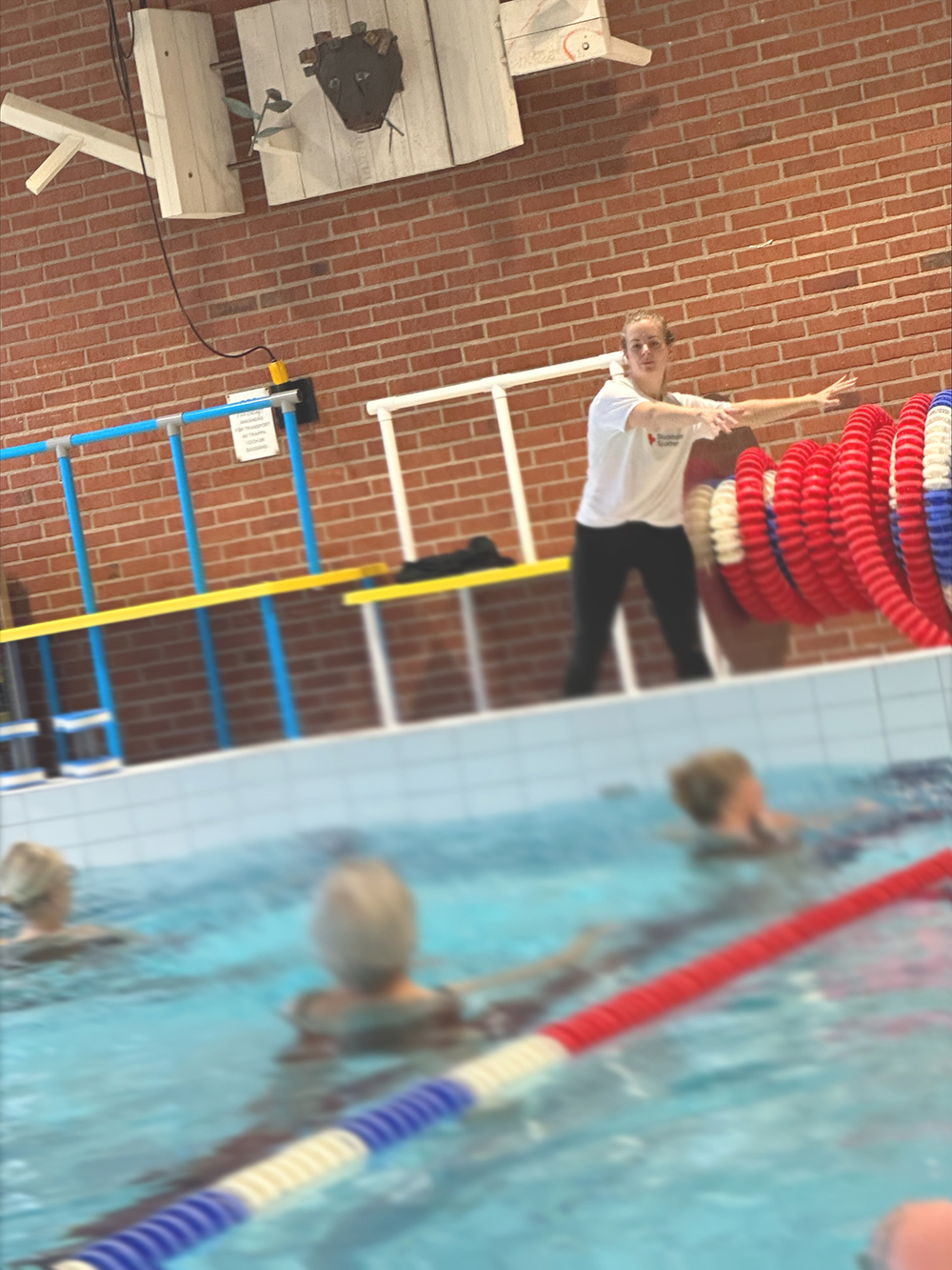 Instruktör leder vattengympa för äldre personer i en simbassäng, väggen är av röd tegel.