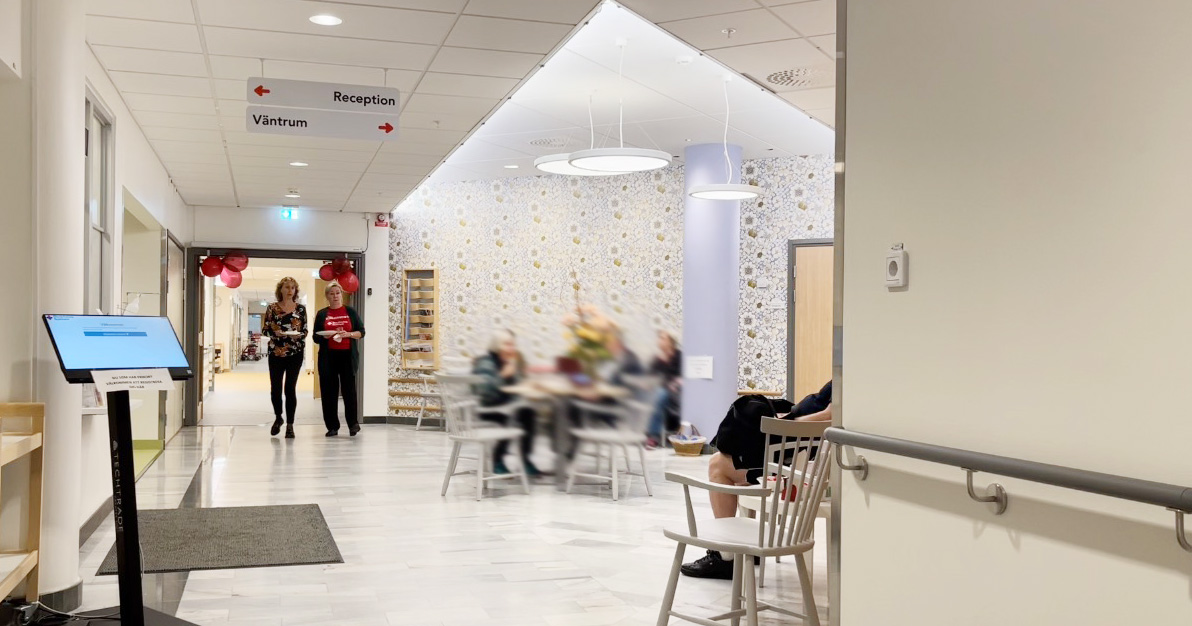 Väntrum på vårdcentral med folk som sitter och väntar, två kvinnor går in, tydliga skyltar till reception och väntrum.