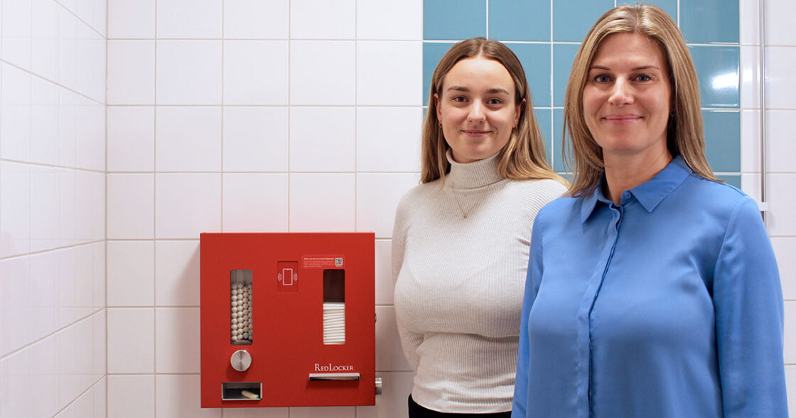 Två kvinnor står bredvid en röd varuautomat för mensskydd på en offentlig toalett med vita kakelväggar.