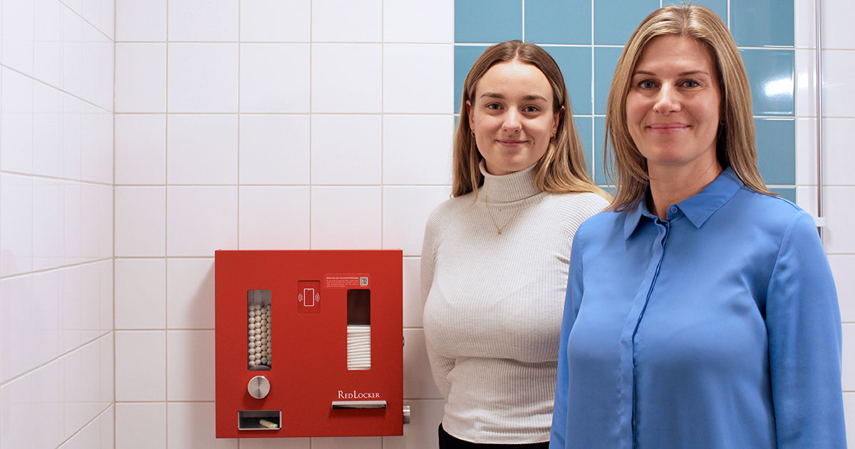 Två kvinnor står bredvid en röd varuautomat för mensskydd på en offentlig toalett med vita kakelväggar.