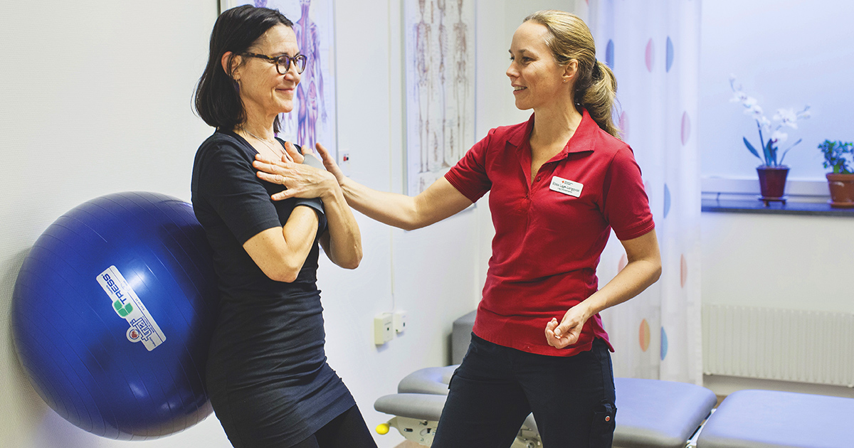 En kvinna får hjälp med träningsövningar av en fysioterapeut med en blå pilatesboll mot väggen.