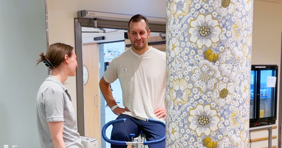 Två personer pratar vid en träningscykel i ett rum med blommönstrad pelare i förgrunden.