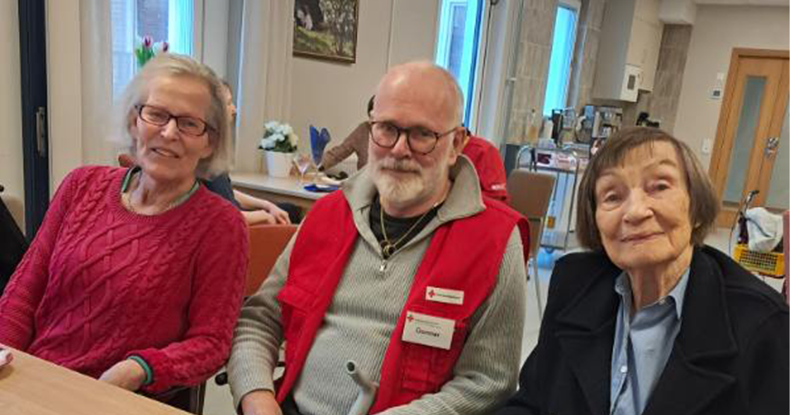 Tre äldre personer sitter vid ett bord i ett ljust rum och ler mot kameran.