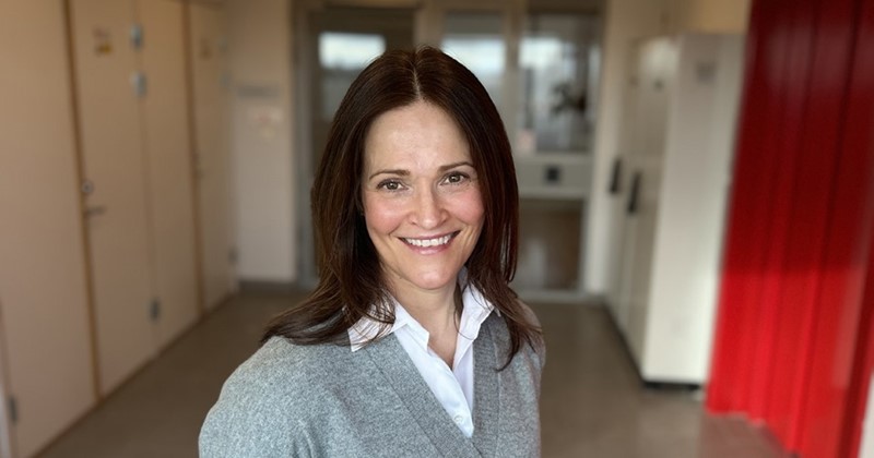 Leende kvinna med mörkt hår i grå tröja står i en korridor med röda gardiner och ljusa väggar.