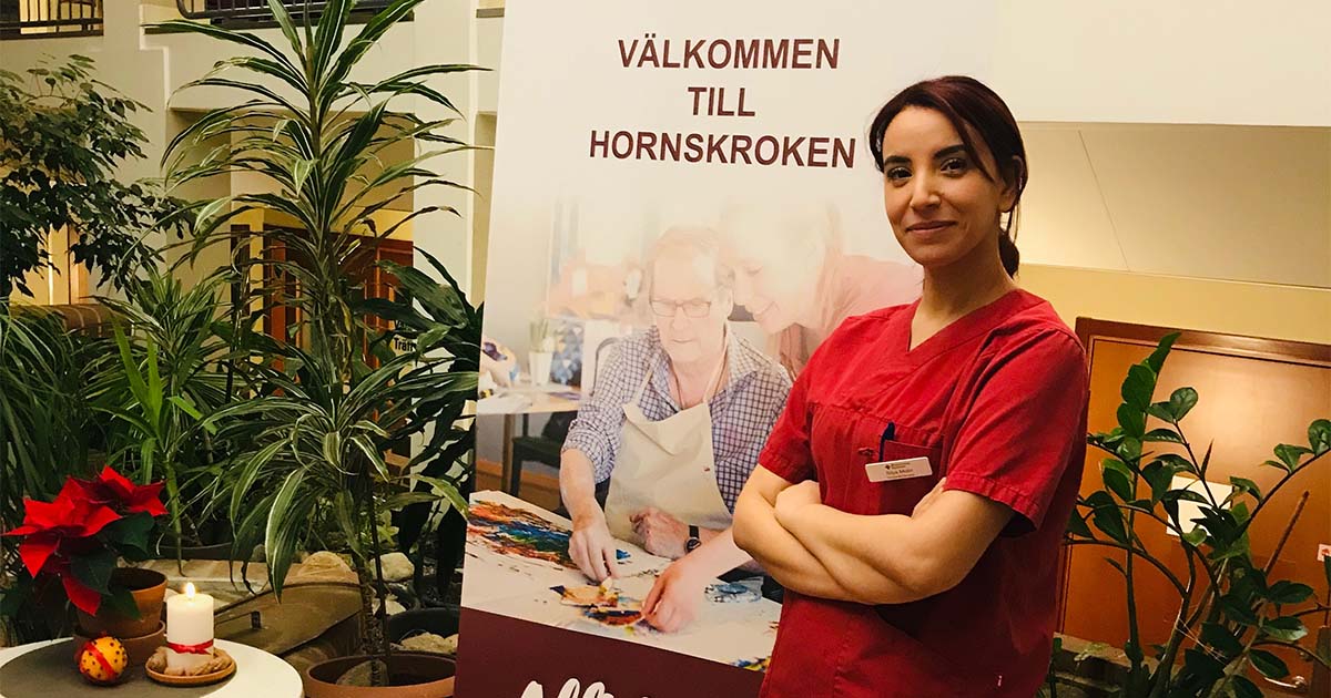Kvinna i röd vårduniform står bredvid en skylt med texten "Välkommen till Hornskroken". Inomhusväxter syns.