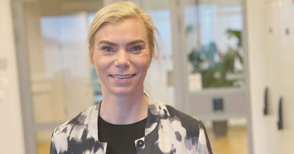 Kvinna med blont hår och svartvit kavaj ler i en ljus inomhusmiljö med kontor i bakgrunden.