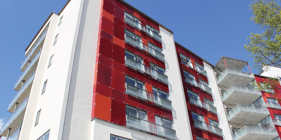 Modernt flerbostadshus med vita väggar, röda paneler och balkonger mot blå himmel.