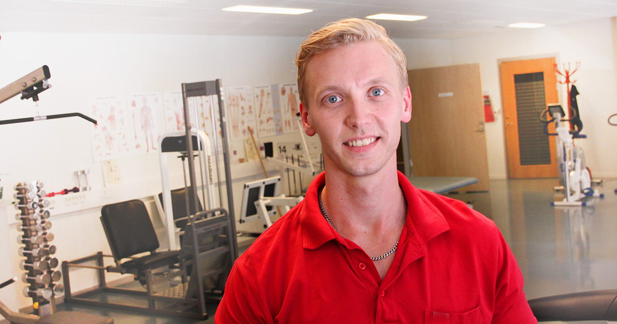 Leende man i röd pikétröja i ett gym eller rehabiliteringsrum med träningsutrustning i bakgrunden.
