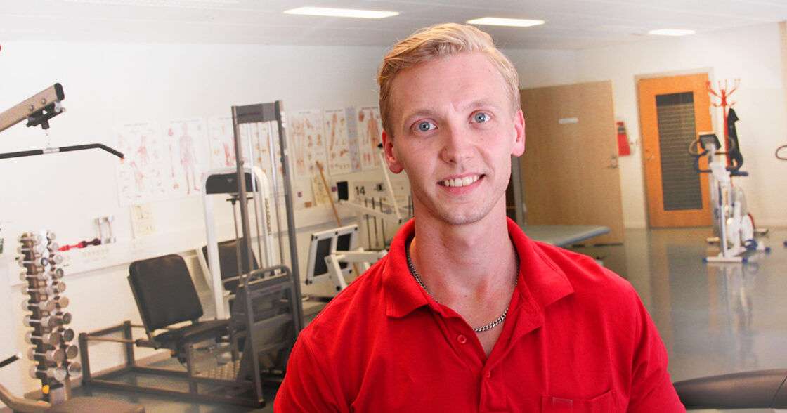Leende person i röd tröja på gym med träningsmaskiner och träningsaffischer på väggen bakom.