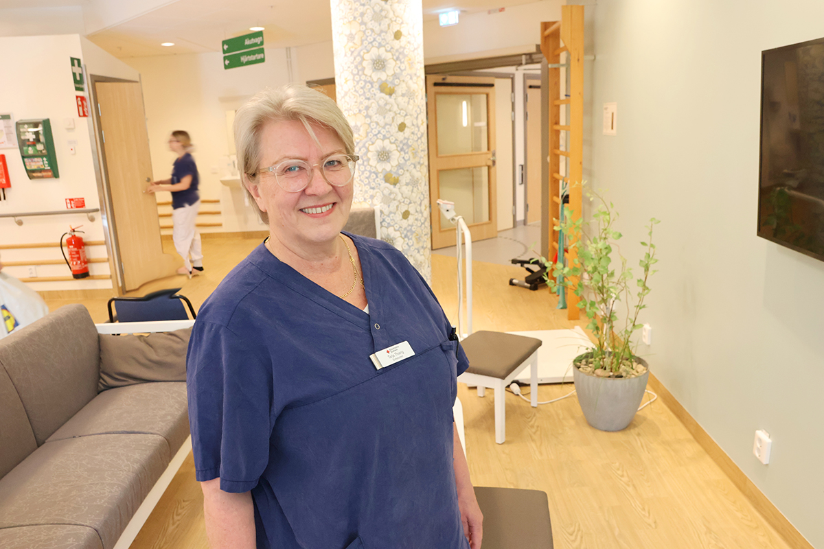 Leende kvinna i blå vårdkläder står i ett ljust väntrum med växter och soffor, sjukvårdsmiljö.