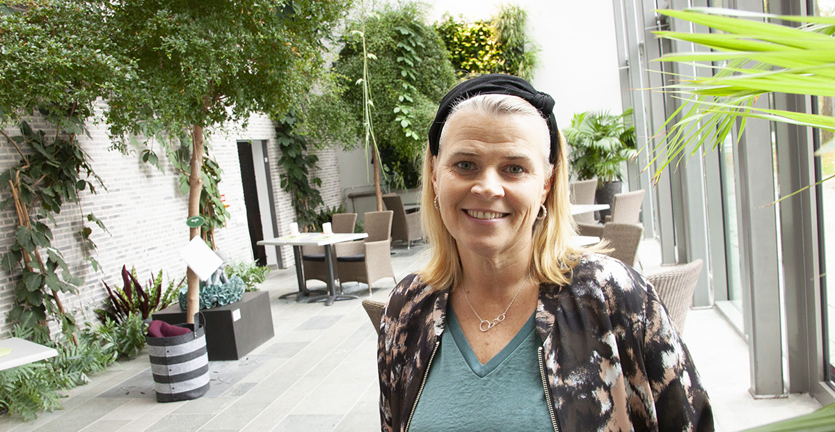 Kvinna med blont hår och hårband står i ett ljust, växtfyllt inomhusutrymme med stolar och bord.