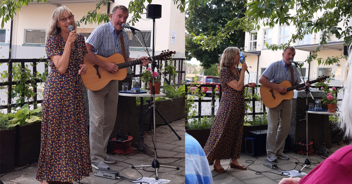 Kvinna sjunger och man spelar gitarr under utomhuskonsert på innergård, omgivna av gröna växter.
